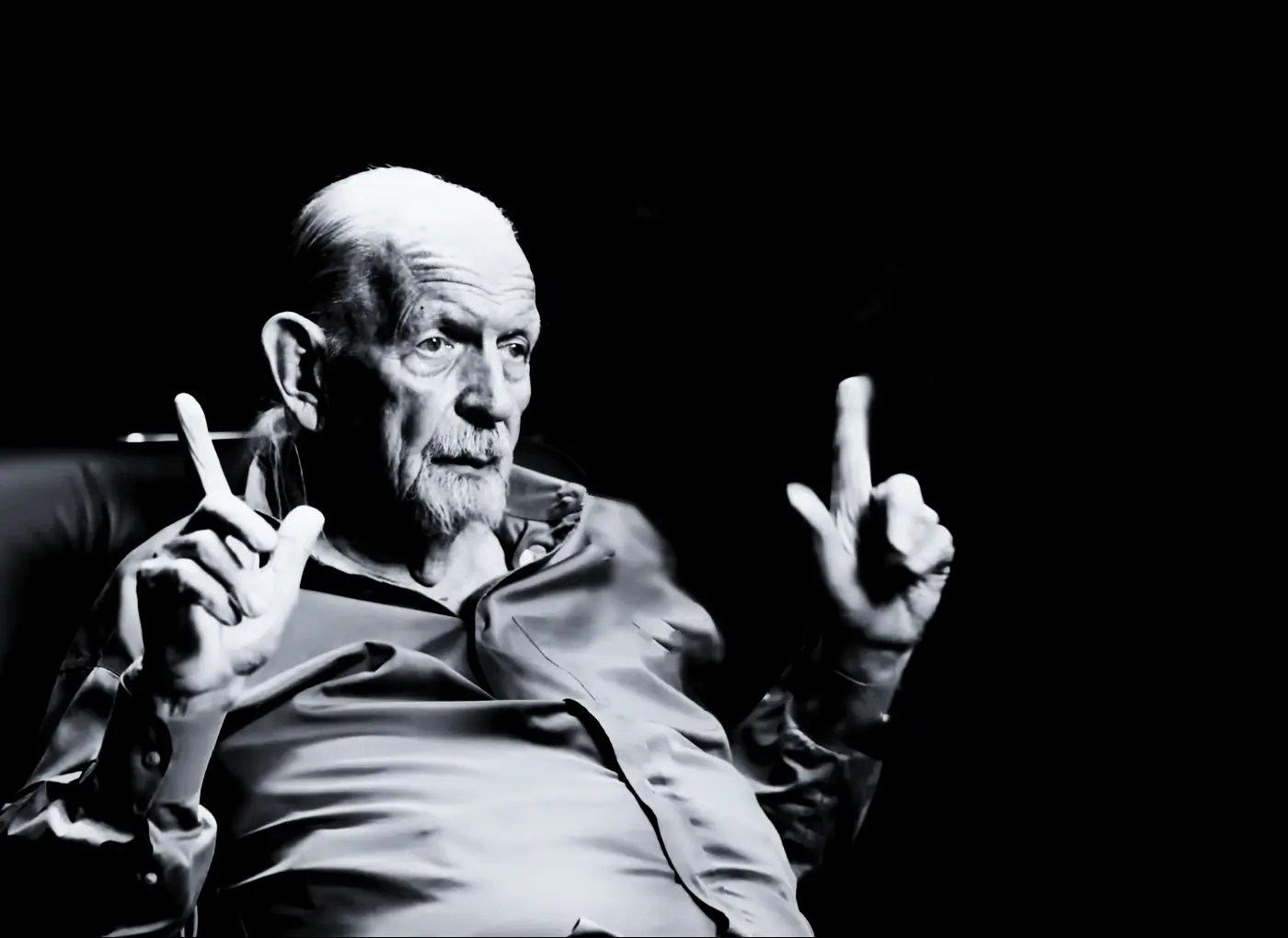 Black-and-white portrait of an elderly man with a white beard, gesturing with both hands, index fingers raised as if signaling.