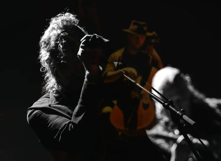 Singer with long hair leaning into a microphone on a dimly lit stage; guitarist in a hat visible in the background.
