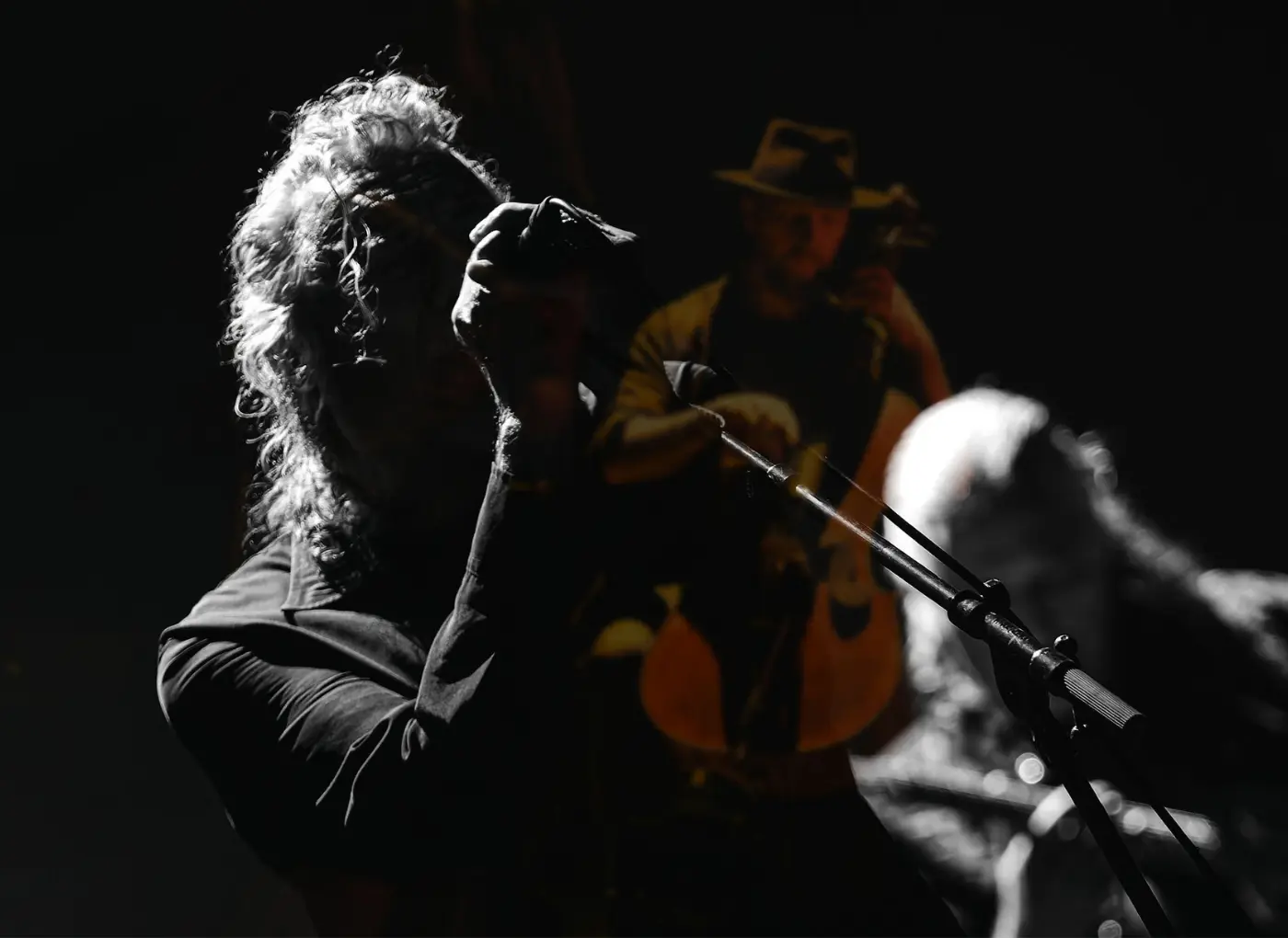 Singer with long hair leaning into a microphone on a dimly lit stage; guitarist in a hat visible in the background.