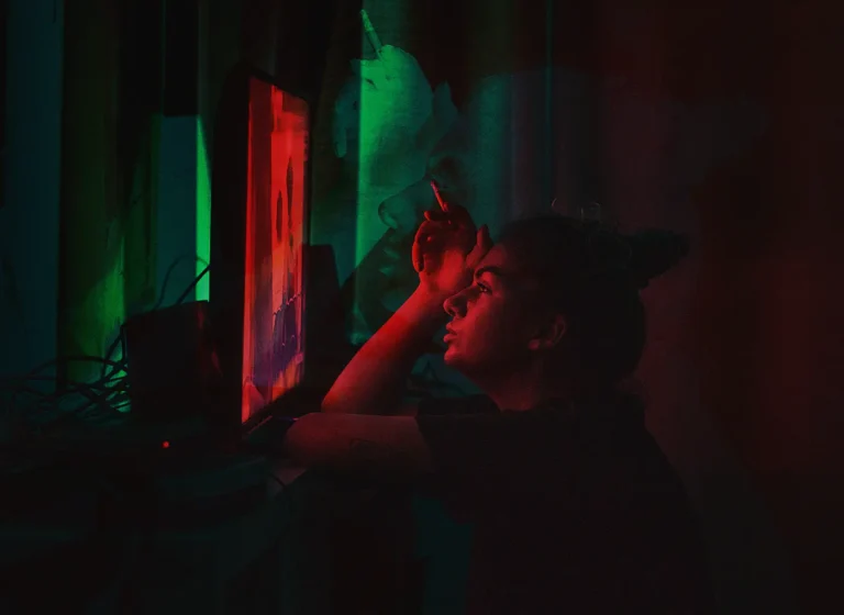 Person sitting in a dark room, illuminated by red and green neon light from a computer monitor, resting their chin on their hand and smoking a cigarette.