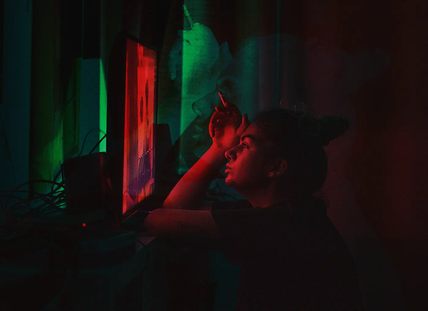 Person sitting in a dark room, illuminated by red and green neon light from a computer monitor, resting their chin on their hand and smoking a cigarette.