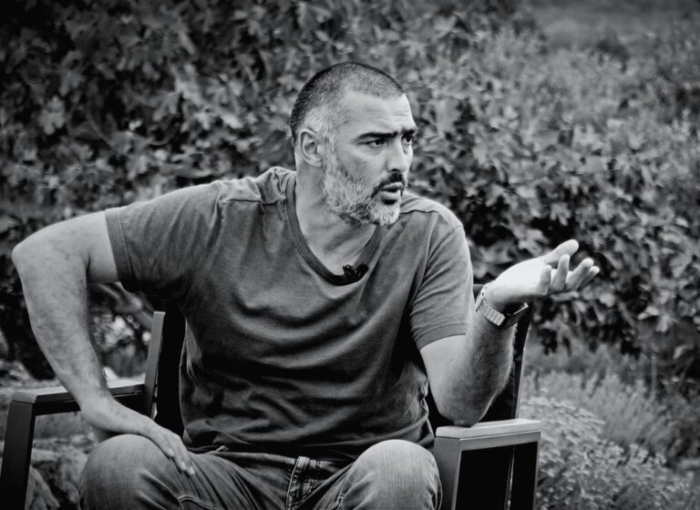 Man with a beard sits outdoors in a chair, gesturing with his right hand as he speaks,