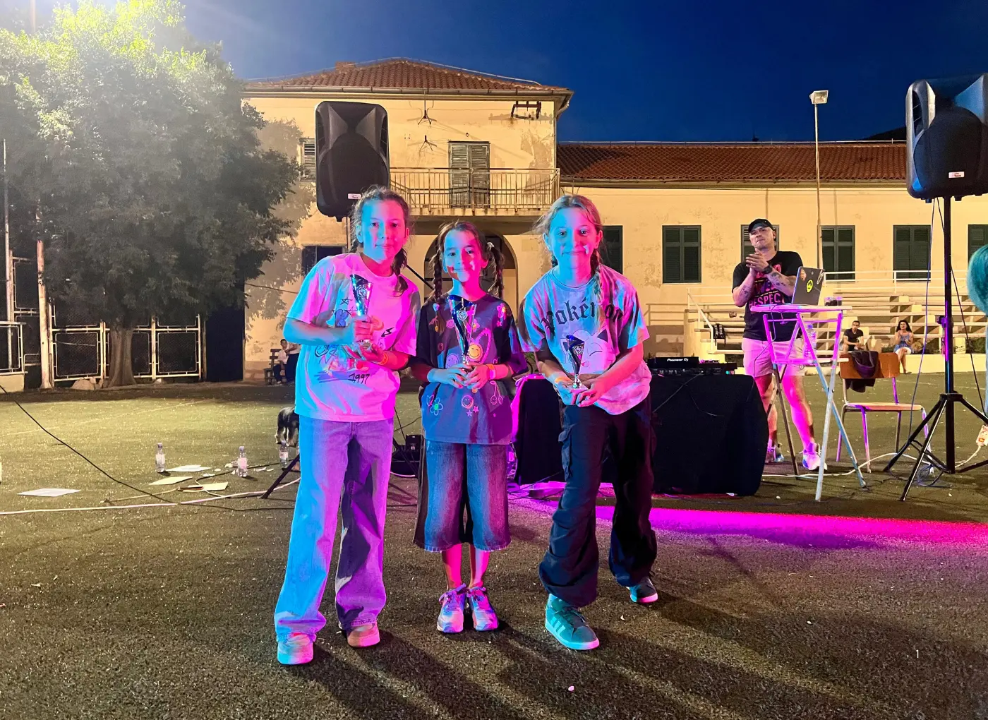 Three young girls pose with trophies on a nighttime outdoor stage, with a DJ and equipment in the background.