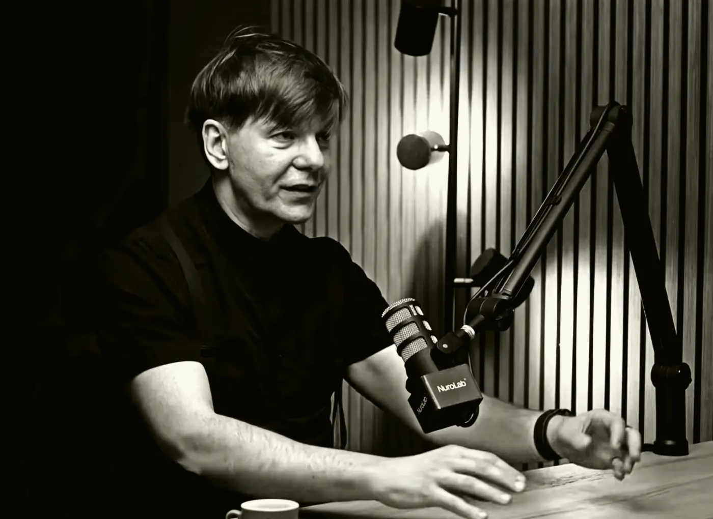 Man in a black shirt speaks into a studio microphone at a recording booth with acoustic panels behind him.—Informative image for podcast/recording context.