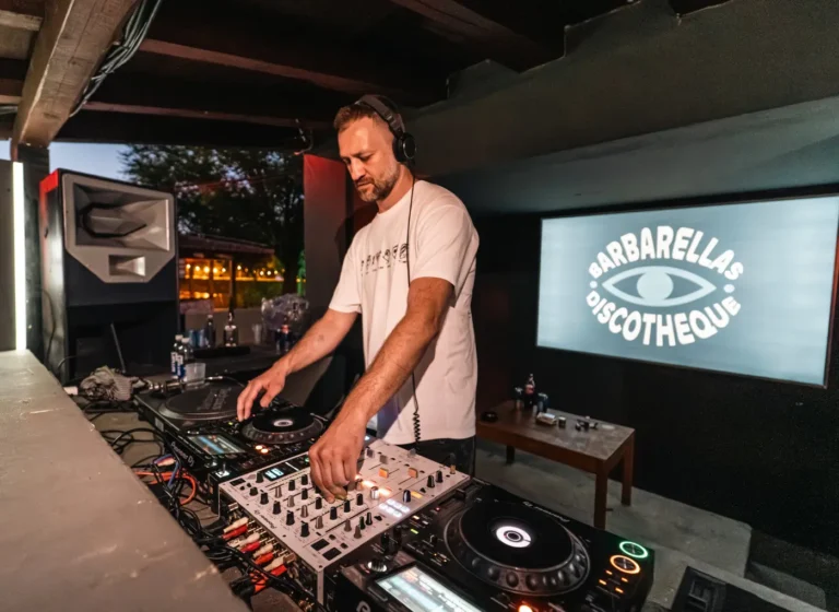 DJ wearing headphones and white shirt performs behind a mixer and turntables at a venue, with a logo screen reading BARBARELLAS DISCOTHÈQUE in the background