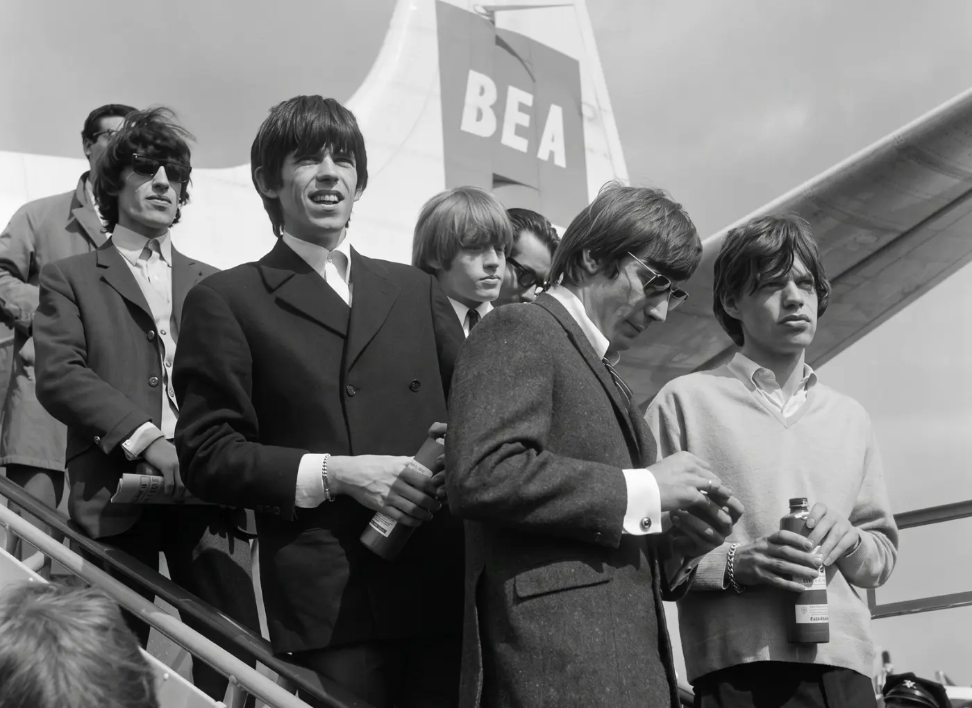Group of young men in suits on airplane stairs, holding bottles, with a BEA tail in the background.