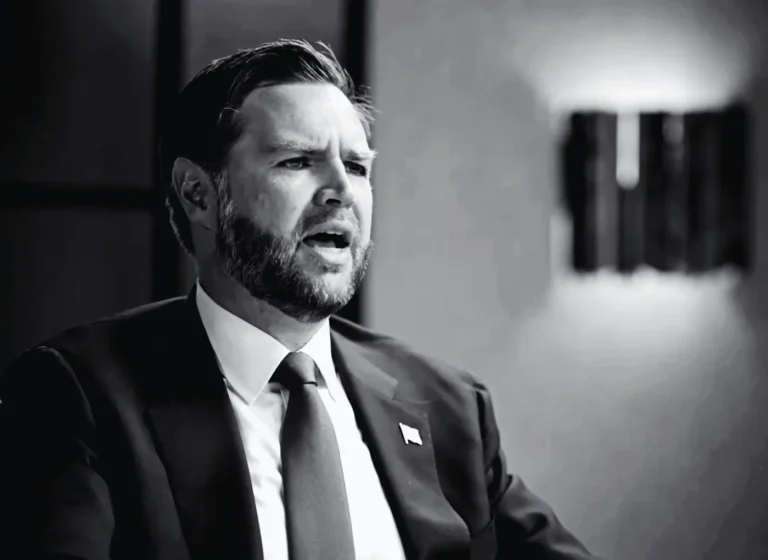 Businessman in a dark suit and tie speaks with a serious expression in a grayscale photo.