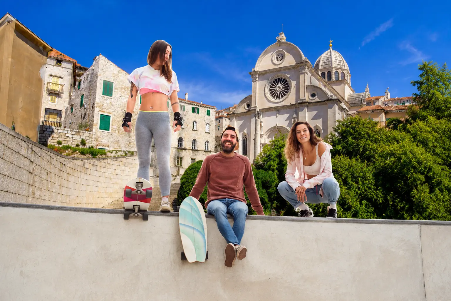 Three friends with skateboards on a concrete ramp in a sunny town square, with a church and stone buildings in the background.