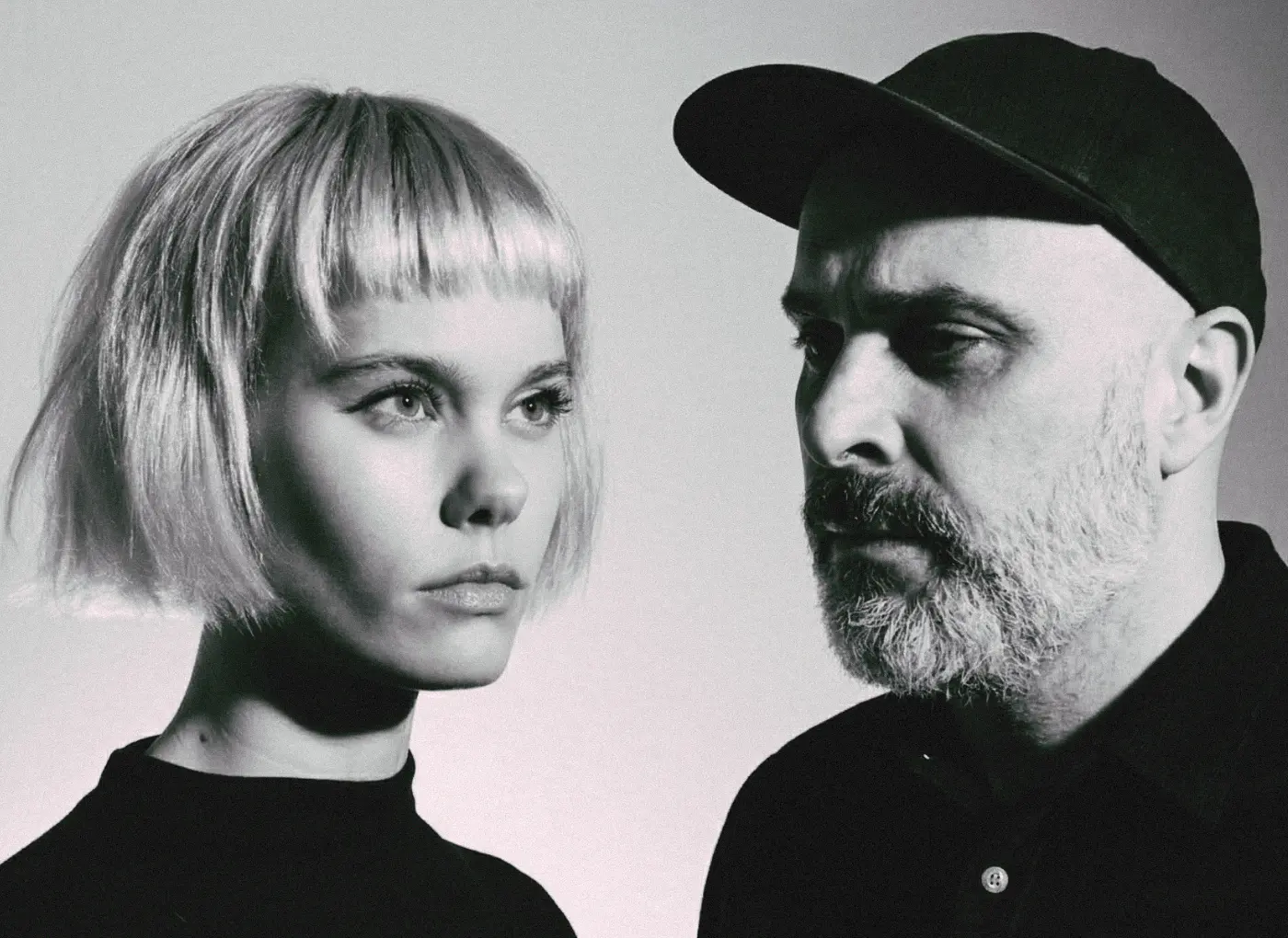 Black-and-white portrait of a woman with a short blonde bob and a bearded man wearing a cap facing each other in profile.