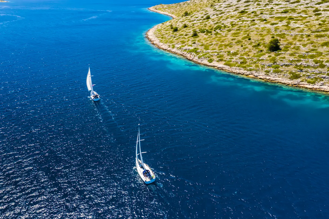 Aerial view of two sailboats sailing along a rocky, green coastline with turquoise shallows and deep blue open sea nearby.