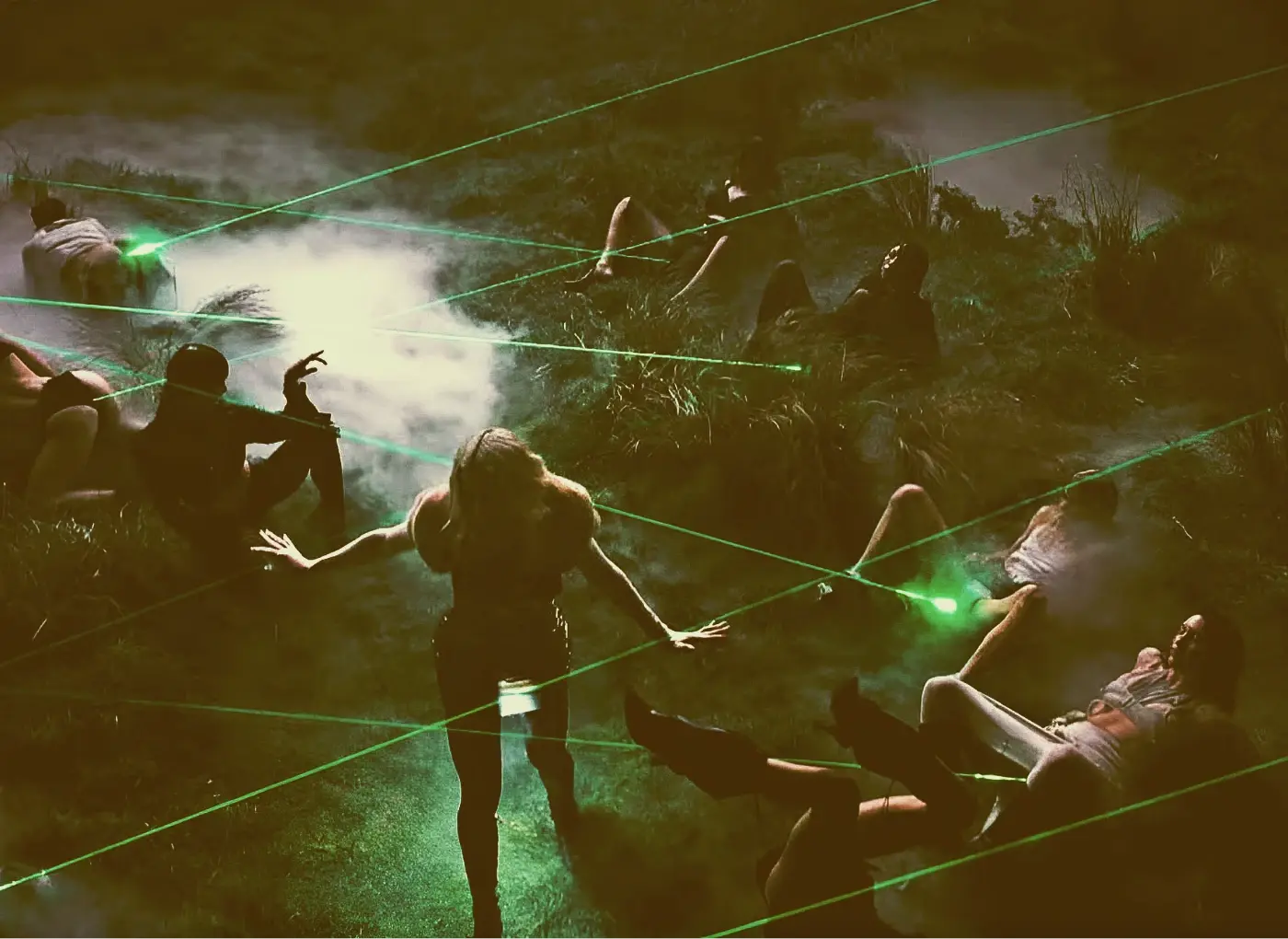 People outdoors at night interacting with green laser beams and fog, silhouetted among tall grass and bushes.