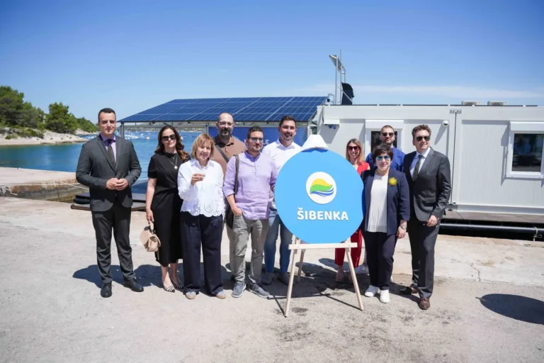 Group of ten adults in business attire posing outdoors beside a blue sign reading ŠIBENKA, with a waterfront and solar-panel building in the background.