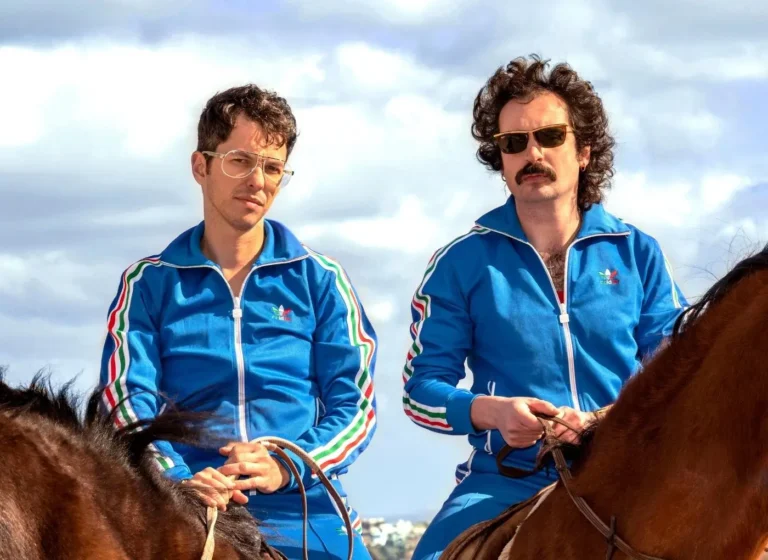 Two men in matching blue tracksuits ride brown horses outdoors on a cloudy day, looking ahead.
