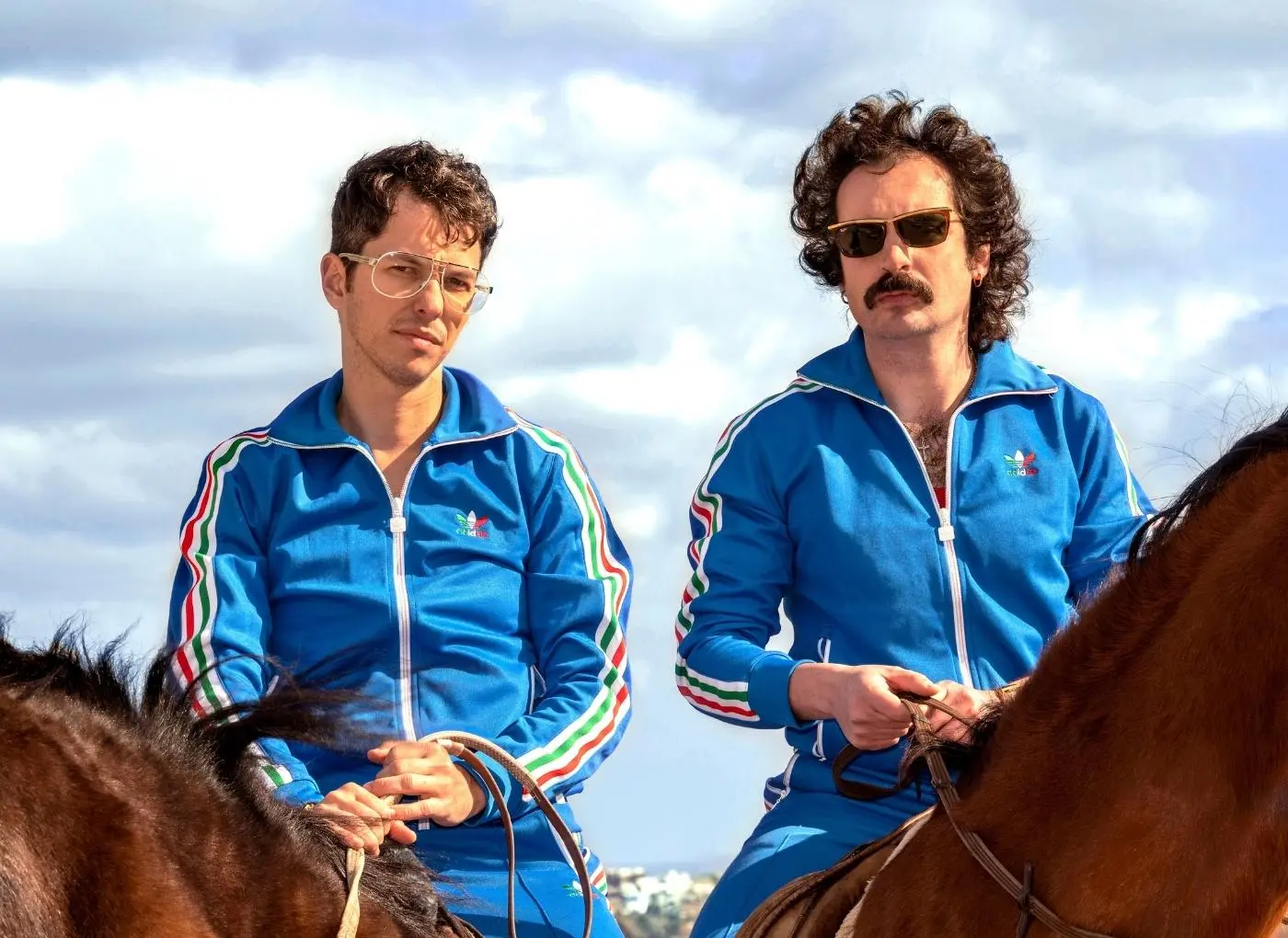 Two men in matching blue tracksuits ride brown horses outdoors on a cloudy day, looking ahead.