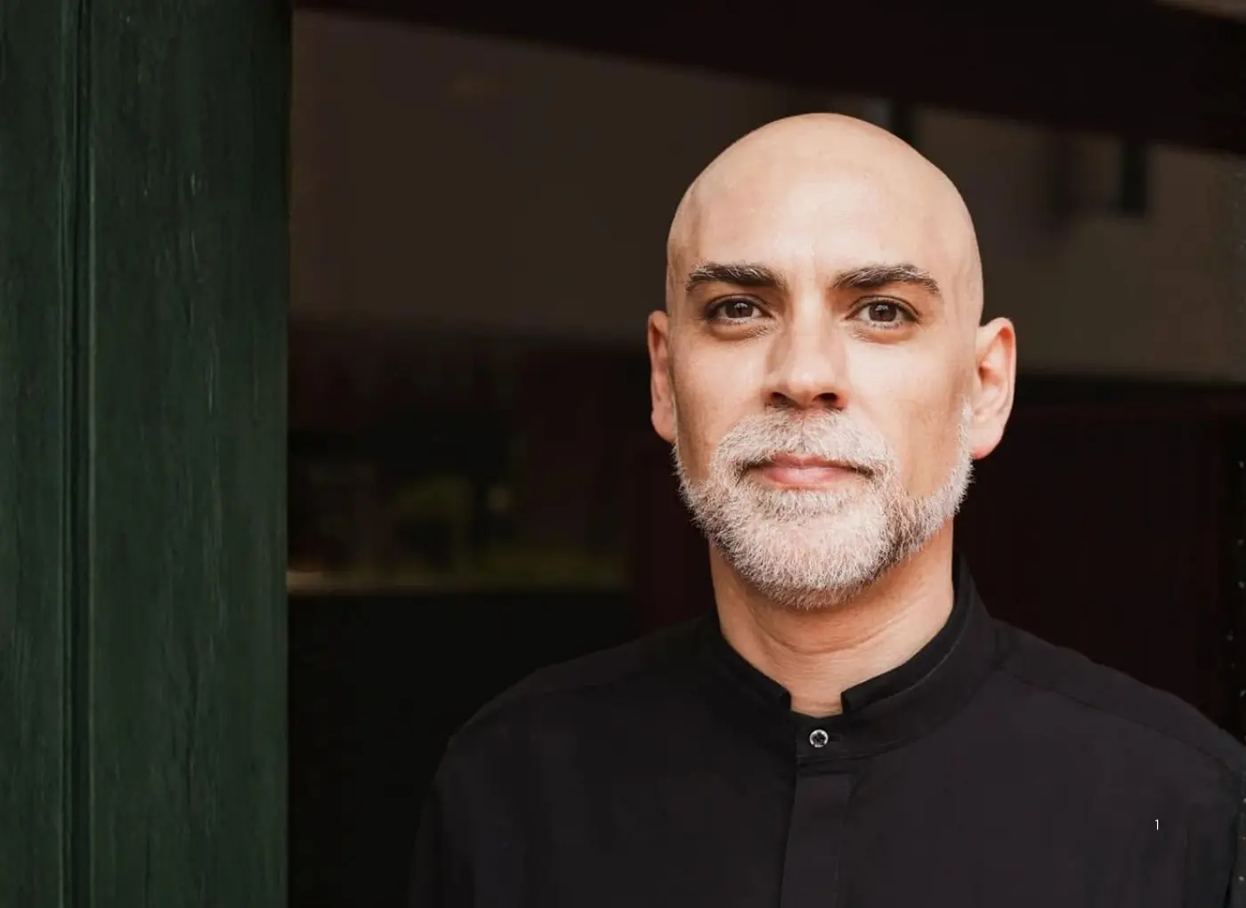 Portrait of a bald, gray-bearded man in a black shirt standing beside a dark doorway or wall, looking at the camera.