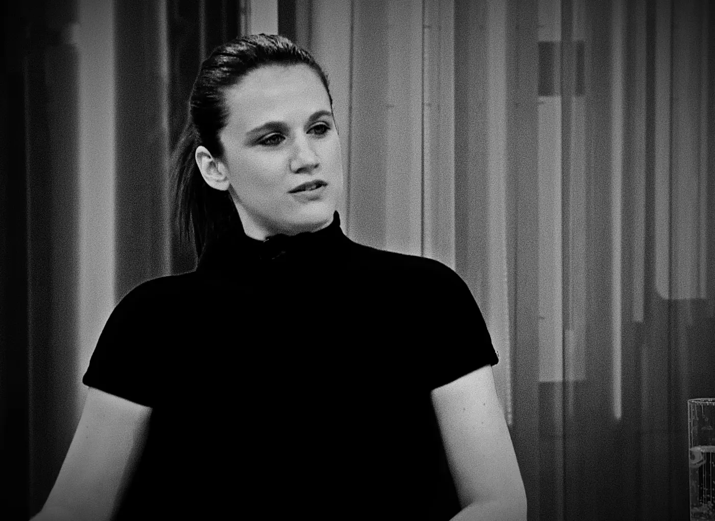 Black-and-white portrait of a woman in a black turtleneck looking to the left, with vertical blinds in the background.
