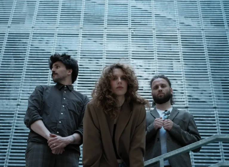 Three stylish adults pose in front of a blue-gray, slatted metal wall: a man on the left, a curly-haired woman in the center, and a bearded man on the right in a blazer.