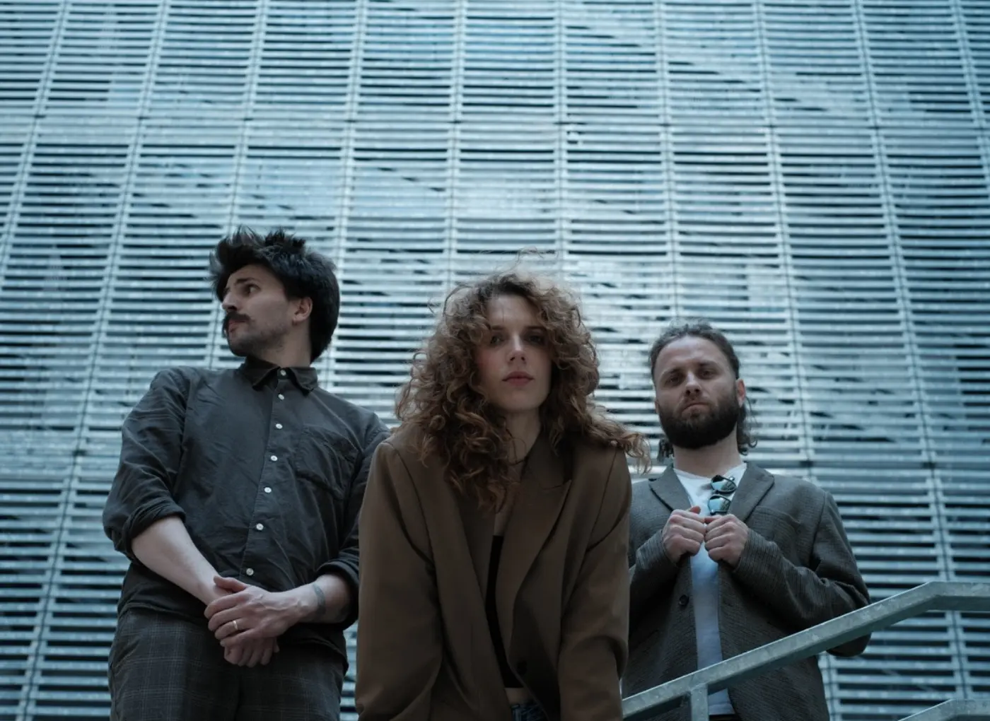 Three stylish adults pose in front of a blue-gray, slatted metal wall: a man on the left, a curly-haired woman in the center, and a bearded man on the right in a blazer.