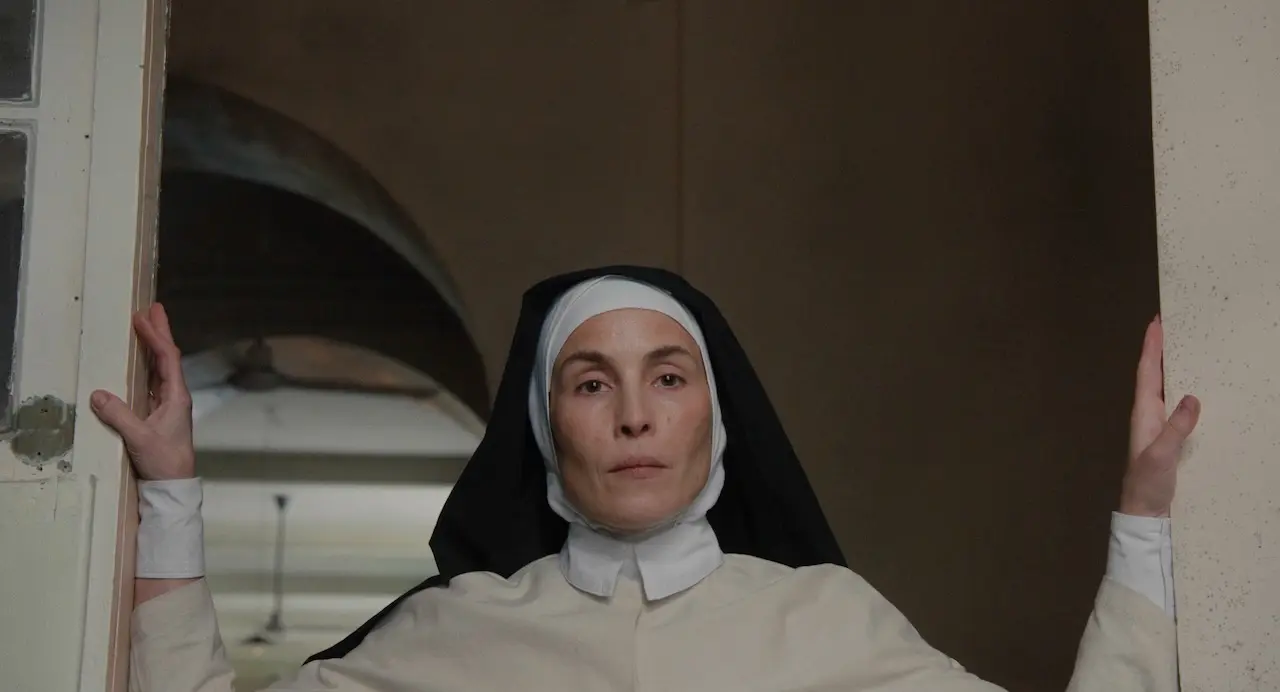 A nun in a white habit and black veil stands in a doorway, hands on the door frame and a calm, serious expression.