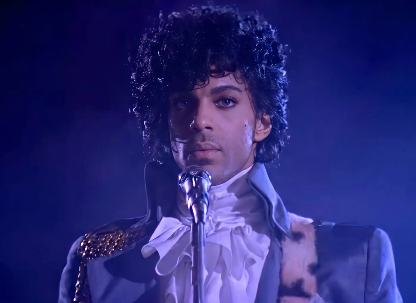 Male performer with dark, curly hair in a blue stage jacket with gold shoulder details, singing into a vintage microphone under blue stage lighting.