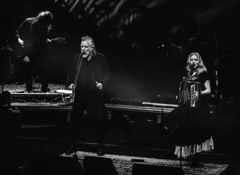 Black-and-white stage scene with a male singer at center mic and a female singer playing accordion on the right, during a live concert.