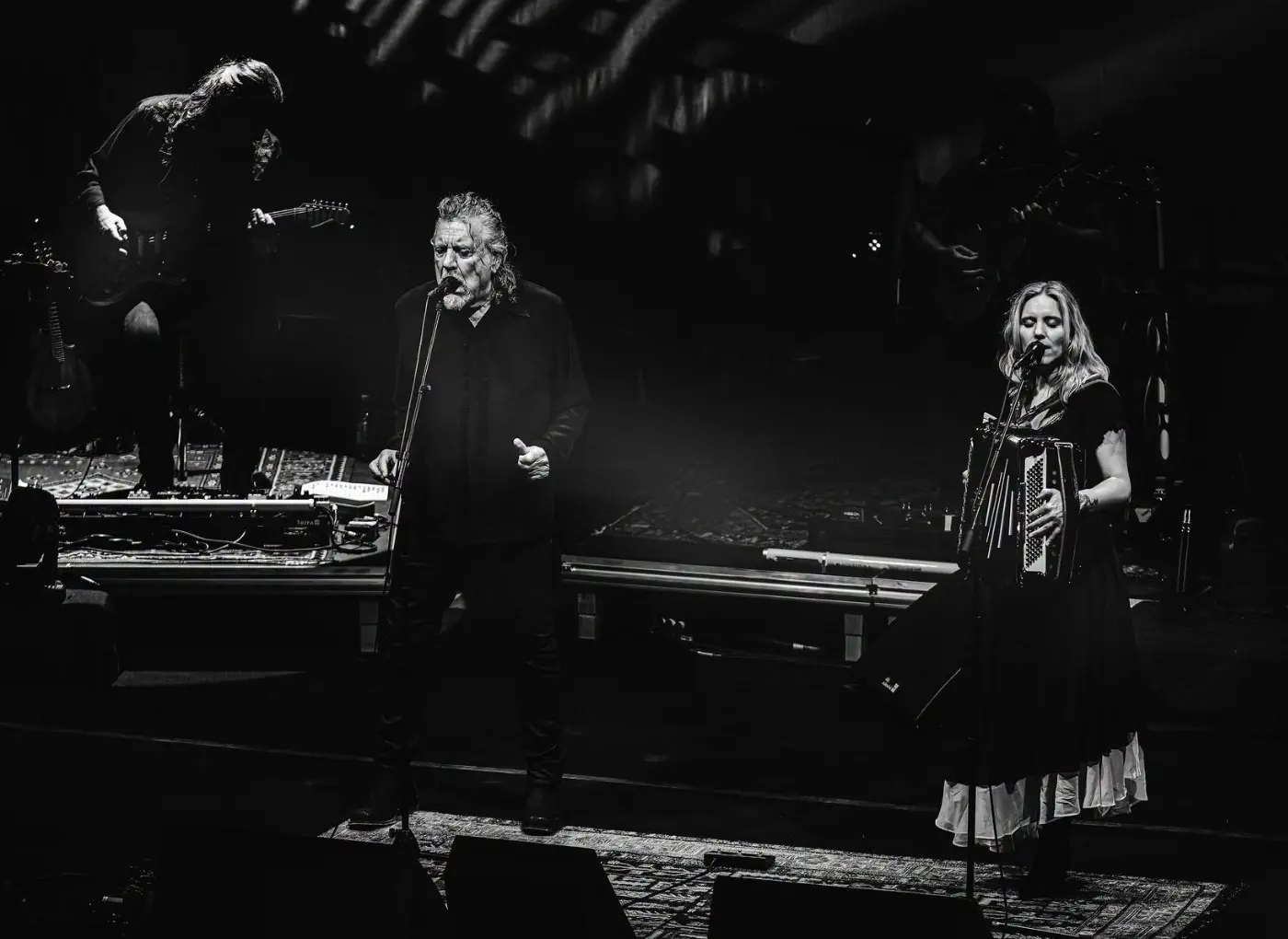 Black-and-white stage scene with a male singer at center mic and a female singer playing accordion on the right, during a live concert.