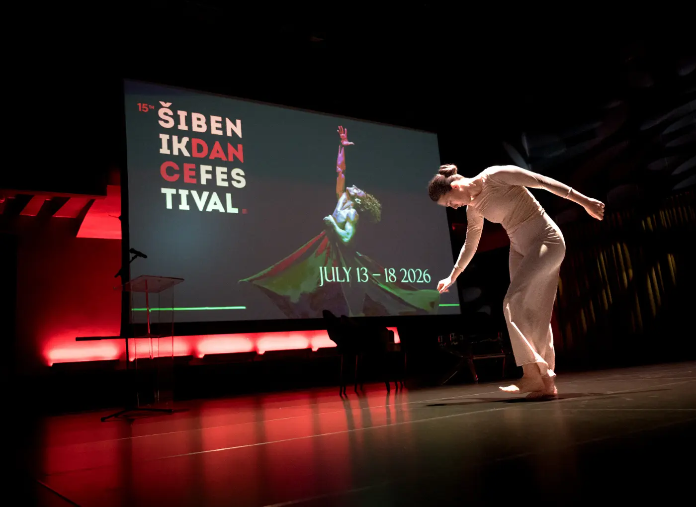 Dancer in beige dress bends forward on stage during the Šibenik Dance Festival; large screen shows festival name and dates (July 13–18, 2026).