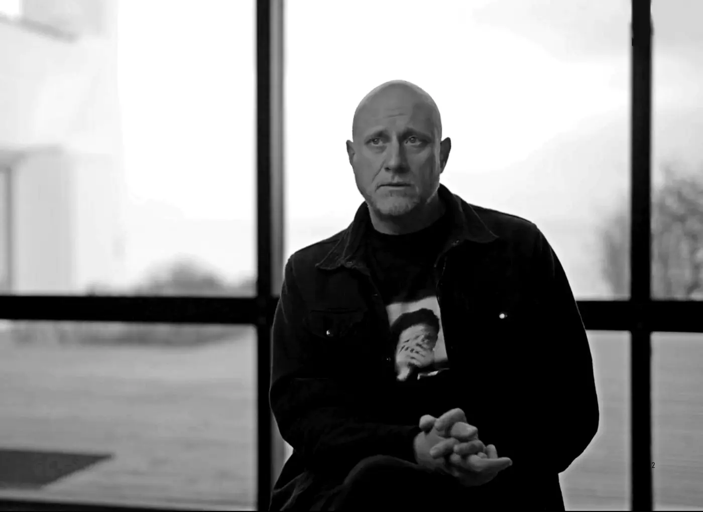 Black-and-white photo of a bald man in a dark jacket sitting with hands clasped, looking thoughtful in front of large windows.