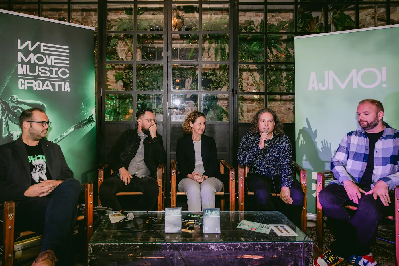 Five panelists seated in a row during a discussion, microphones on the glass coffee table, with green banners reading 'We Move Music Croatia' and 'AJMO!' in the background.