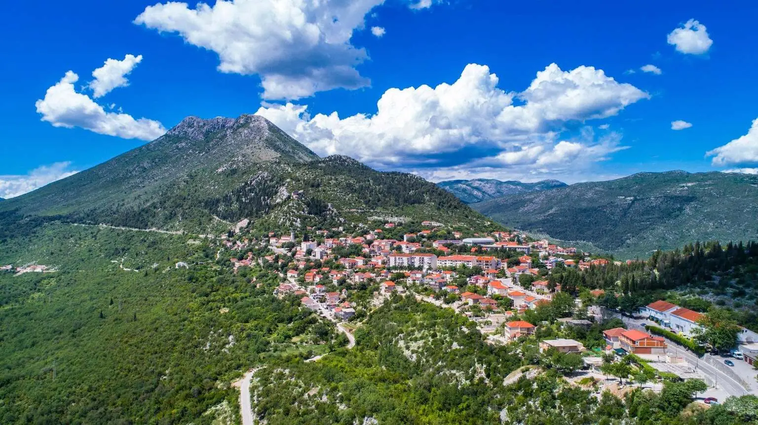 A hillside village with red-roofed houses scattered along a winding road at the base of a green mountain under a bright blue sky with fluffy white clouds.