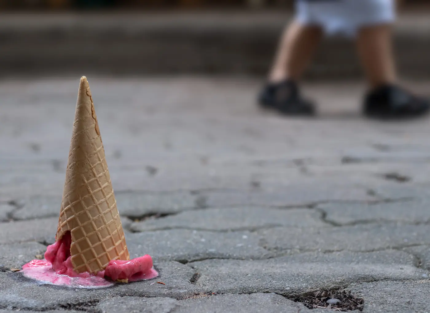 Melting ice cream cone on cracked pavement with a blurred person walking in the background.