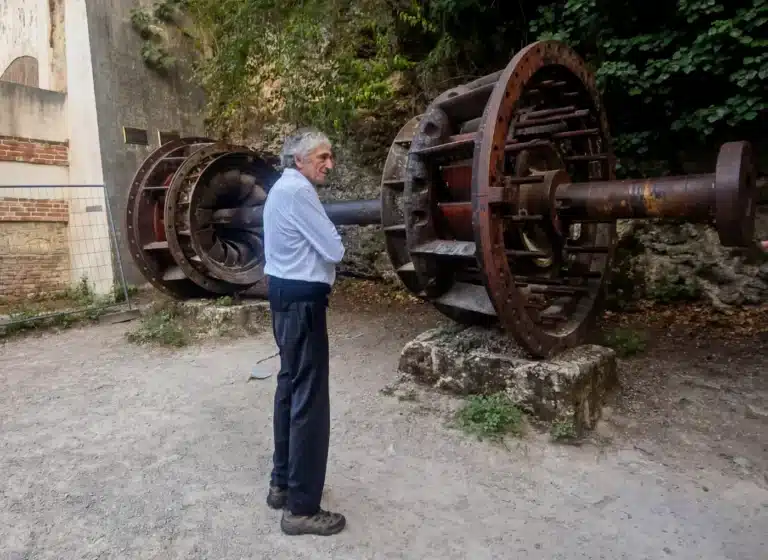 Older man in a light blue shirt and dark trousers stands with arms crossed, facing a large rusted industrial wheel on a shaft outdoors.