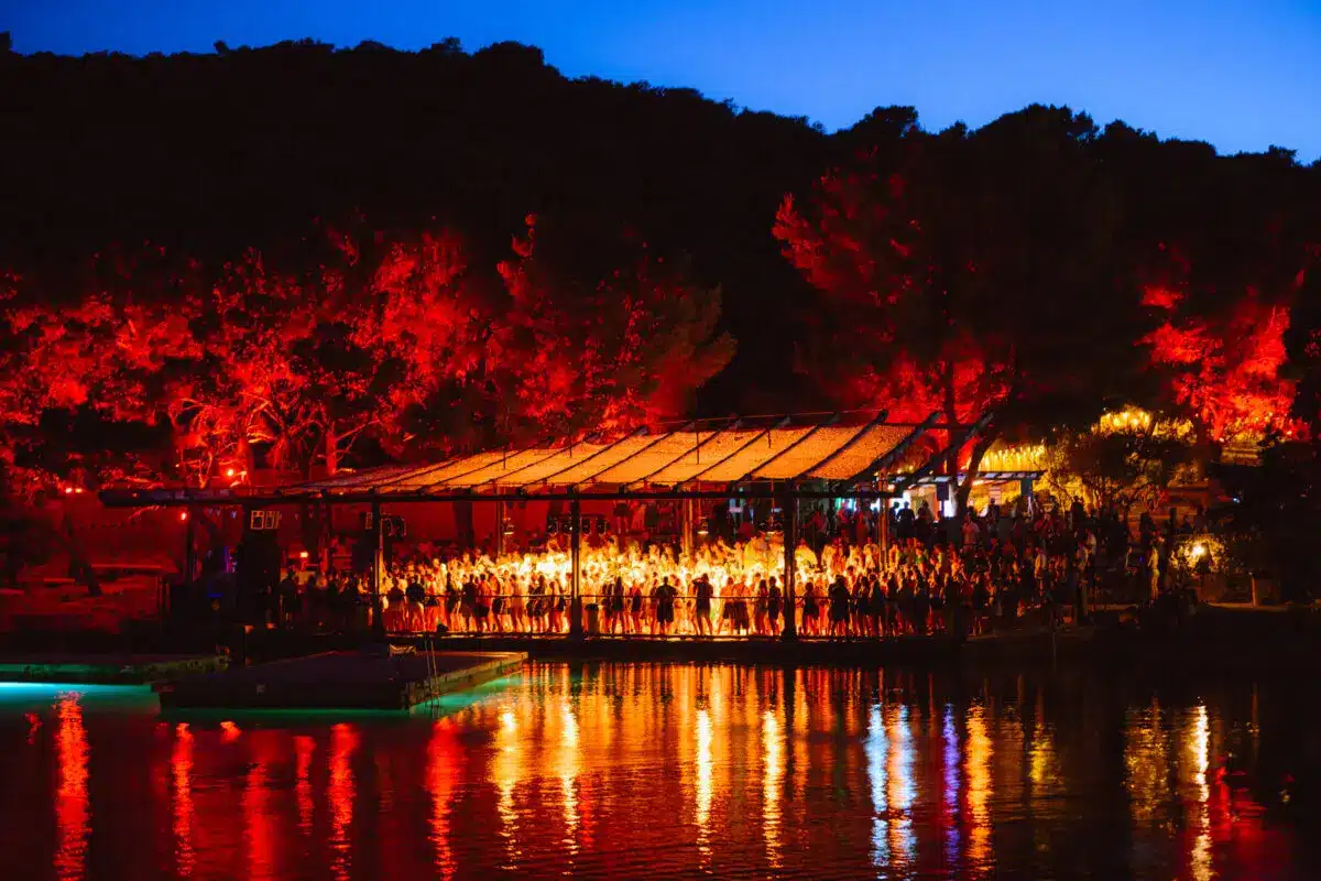 Night scene of a crowded dockside pavilion lit with warm orange lights, red trees in the background, reflections on the water.