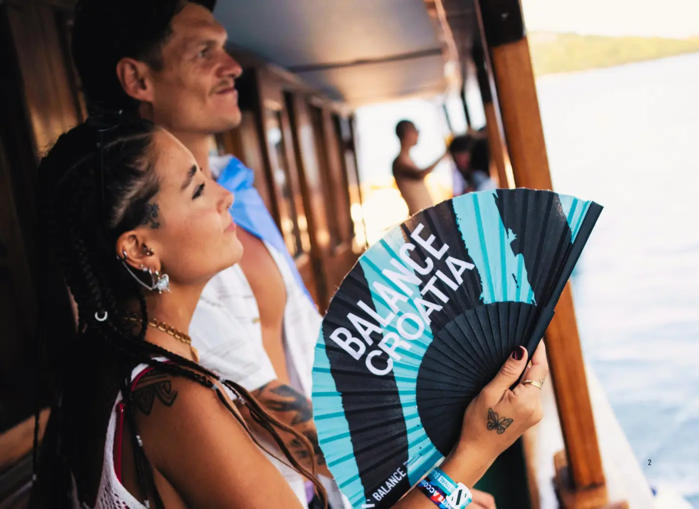 Woman on a wooden boat, holding a blue and black folding fan with the words BALANCE CROATIA, near the water with sunlight in the background.