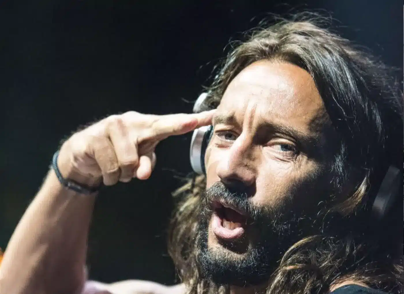 Bearded man with long hair wearing headphones, touching his temple with a finger as if thinking.