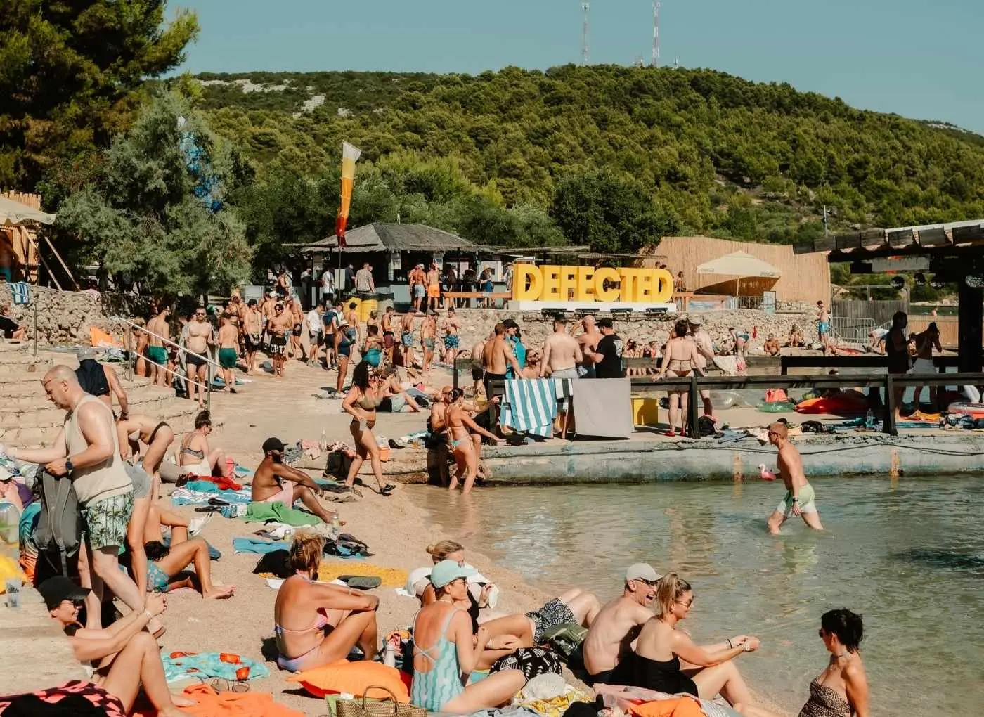 Crowded beach scene with sunbathers along the shore and a large yellow 'DEFECTED' sign in the background.