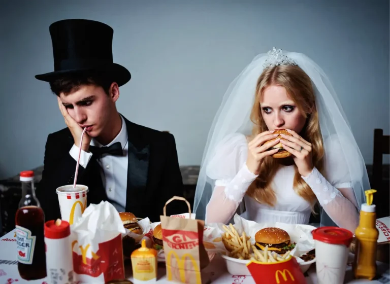 Bride in a veil and gown and groom in a tuxedo sit at a table of fast food, both looking weary; the groom drinks from a straw, the bride bites a burger.