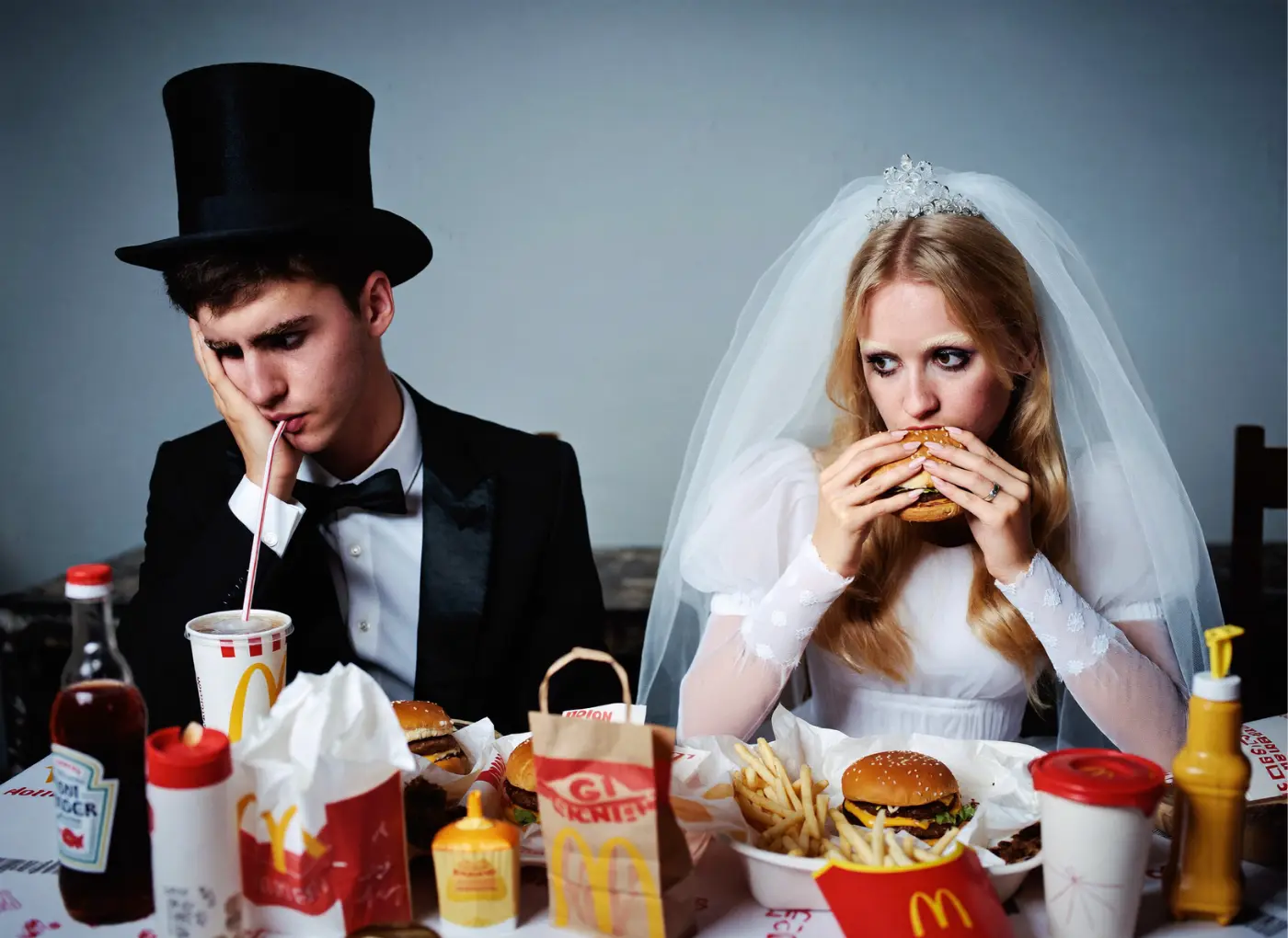 Bride in a veil and gown and groom in a tuxedo sit at a table of fast food, both looking weary; the groom drinks from a straw, the bride bites a burger.
