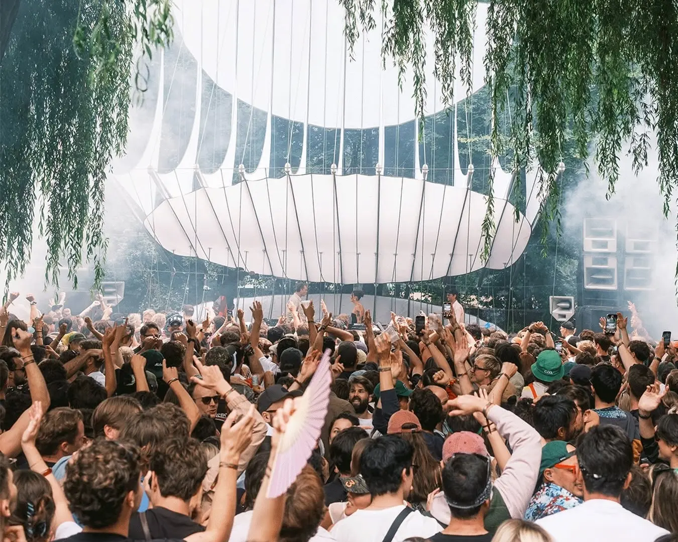 Dense crowd at an outdoor music festival, hands raised toward a large white circular stage with hanging greenery above.