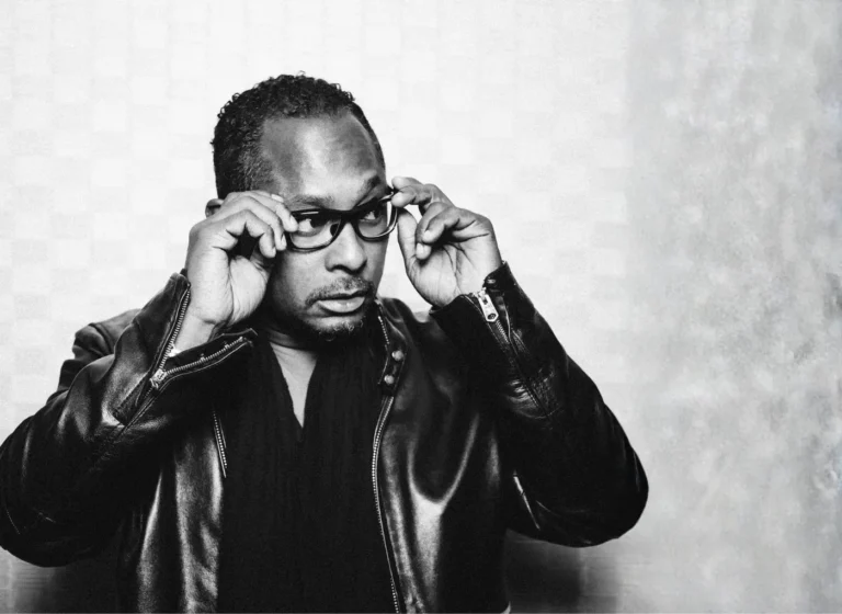 Black-and-white portrait of a man in a leather jacket adjusting his thick-rimmed glasses, looking sideways.