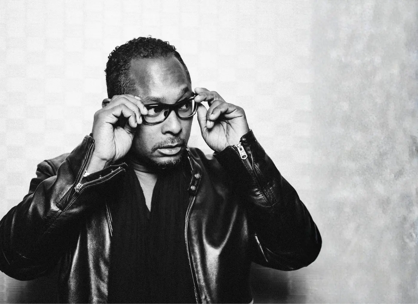Black-and-white portrait of a man in a leather jacket adjusting his thick-rimmed glasses, looking sideways.