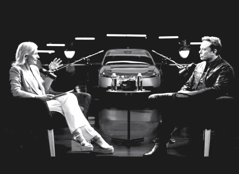 Two hosts in a studio interview, seated across from each other with microphones and a central table between them, a car displayed in the background.