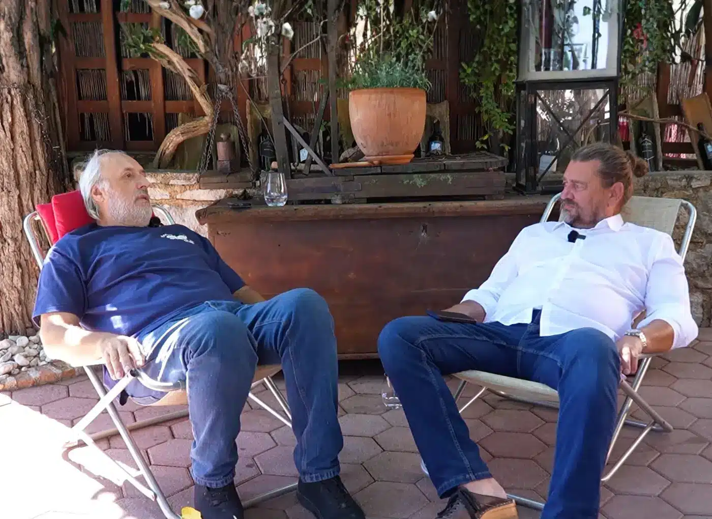 Two men sit in folding chairs on a sunlit outdoor patio, chatting with a glass of water on the table between them.