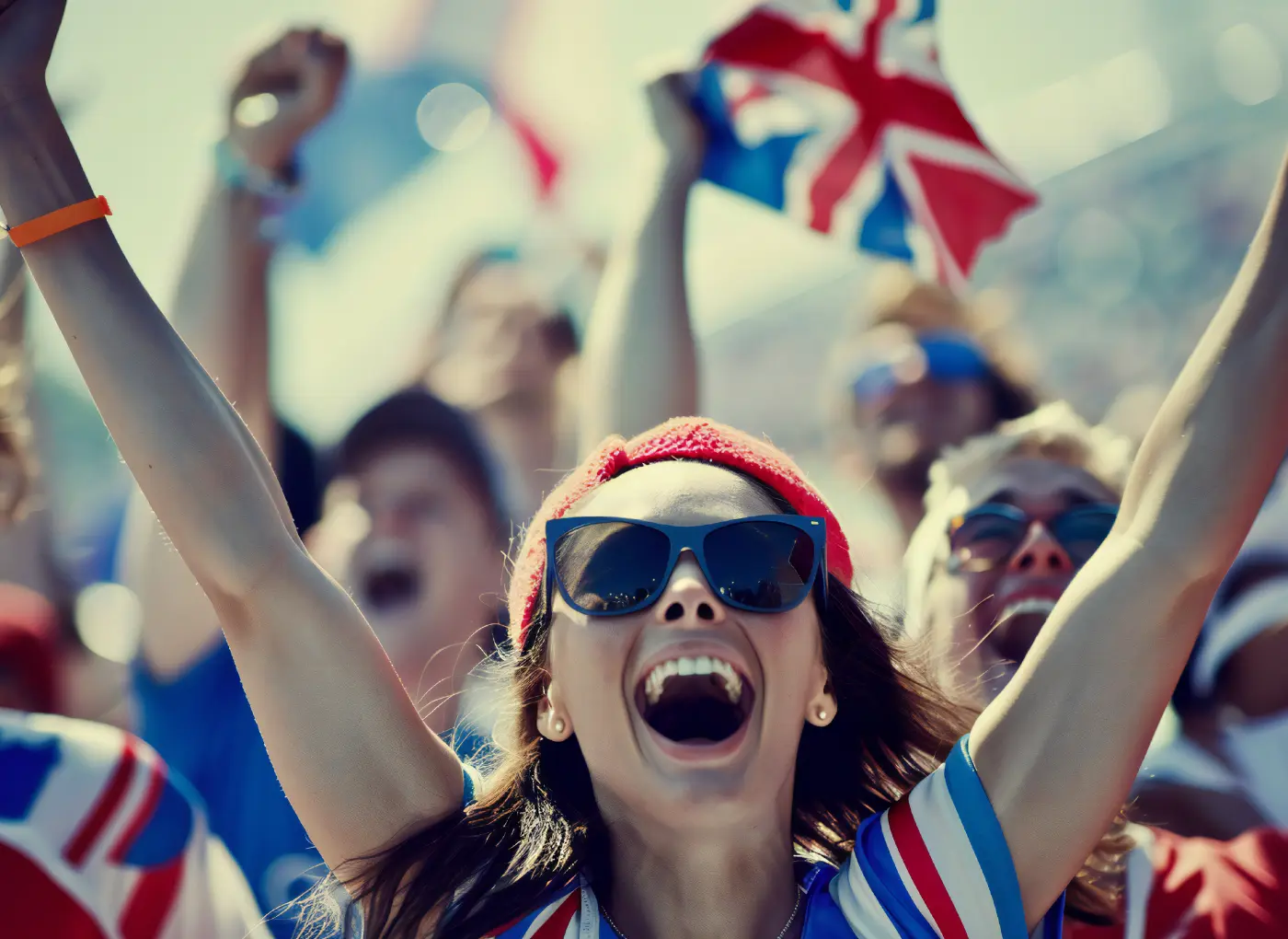 Young woman with a red beanie and sunglasses cheers with arms raised among a cheering crowd at a sports event, flags in the background.