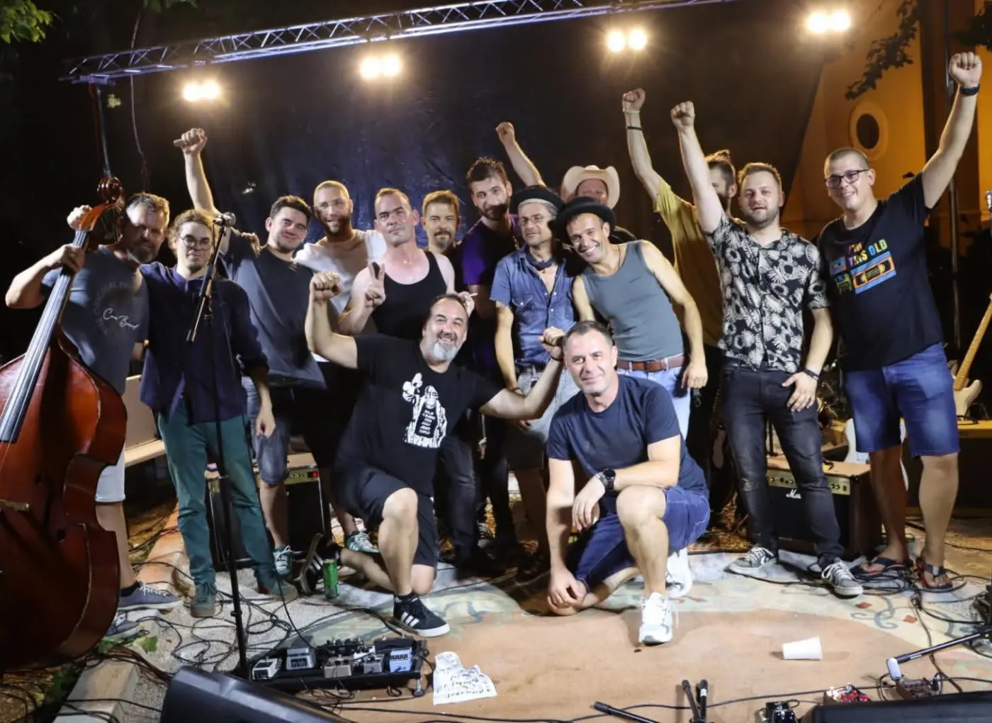 A group of musicians on a stage posing for a photo after a performance, some raising their fists in celebration with instruments visible in the background.