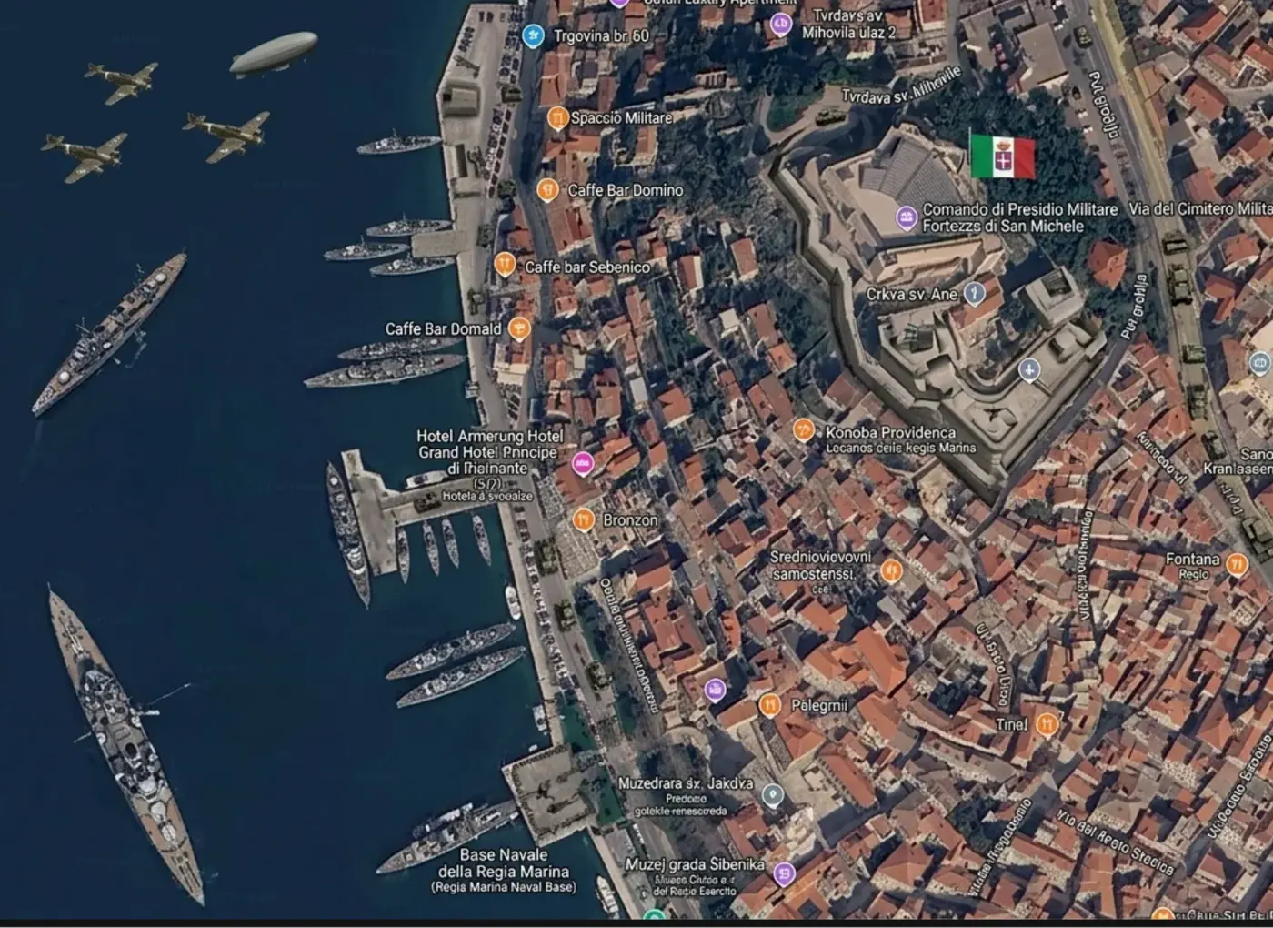 Aerial map of a coastal town with a harbor, red-roofed buildings, and several boats docked along the pier beside dark blue water. Italian flag visible on a hilltop fortress.
