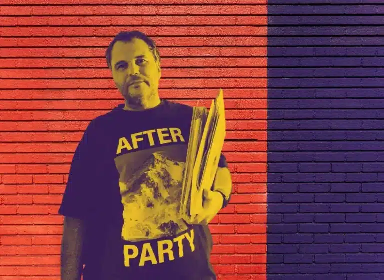 Man standing against a red-and-purple brick wall wearing a black T-shirt that says 'AFTER PARTY' and holding a stack of papers.