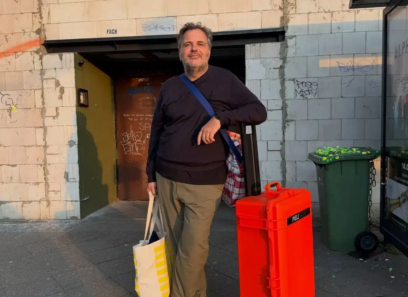 Man stands on a city sidewalk outside a graffiti-covered building, wearing a black shirt and khaki pants, with a plaid bag over his shoulder and a beige striped tote in hand, near an orange wheeled bin.