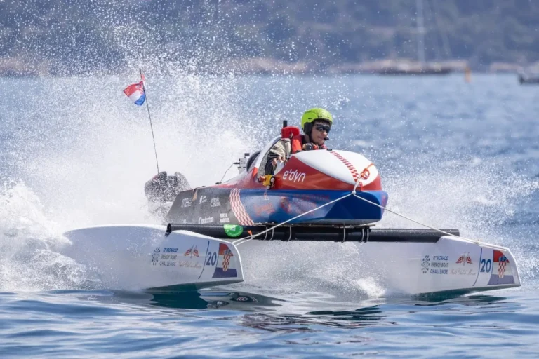 Rider in neon helmet steers a red-and-white speedboat skimming the water, spraying a wide wake during a foiling race.