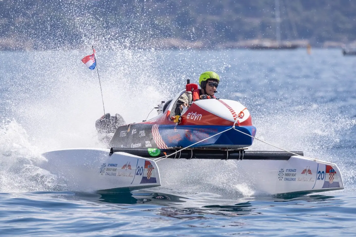 Rider in neon helmet steers a red-and-white speedboat skimming the water, spraying a wide wake during a foiling race.